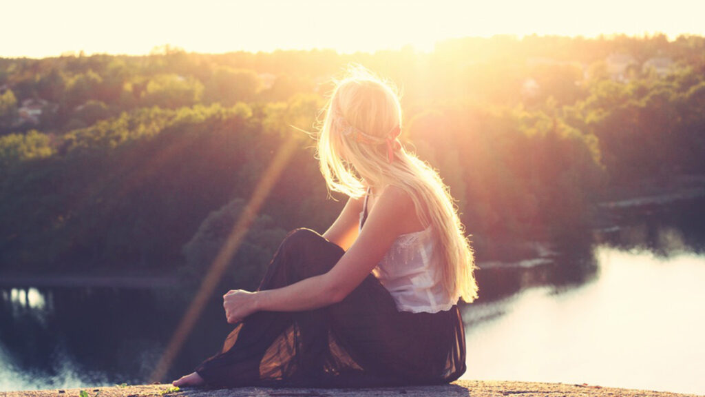 Femme blonde, assise à même le sol sur un point de vue haut, regardant le soleil couchant